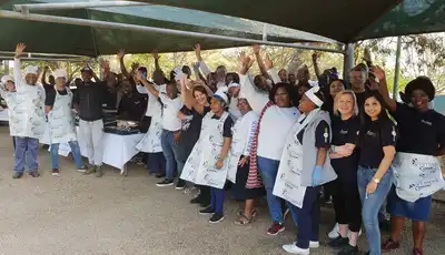 "Group of enthusiastic team members at outdoor event with hands raised, some wearing aprons and Scott Bader branded shirts." [G]