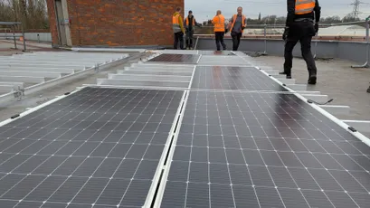 Construction workers installing solar panels on a flat roof, wearing high-visibility orange safety vests. [G]