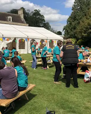 "Outdoor event with attendees in turquoise shirts gathered at wooden picnic tables near a large white marquee tent and historic building." [G]