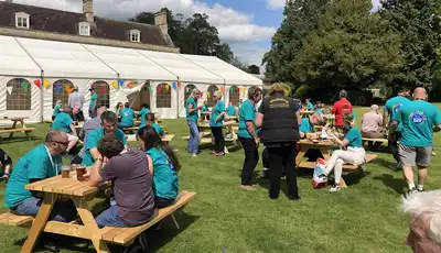 "Outdoor event with attendees in turquoise shirts gathered at wooden picnic tables near a large white marquee tent and historic building." [G]
