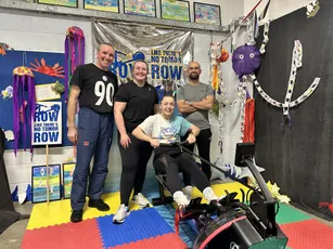 "Group in colorful classroom with exercise equipment and motivational 'ROW' banners on colorful foam mat flooring" [G]
