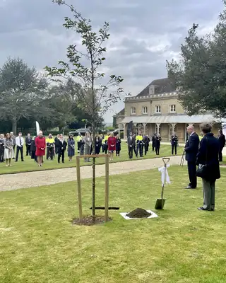 "Tree planting ceremony at Scott Bader grounds with attendees gathered on lawn near historic building" [G]