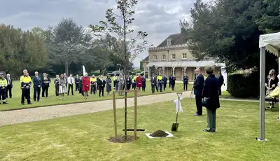 "Tree planting ceremony at Scott Bader grounds with attendees gathered on lawn near historic building" [G]
