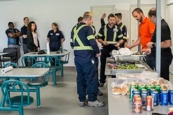 "Industrial workers in safety uniforms gathering for a meal in a cafeteria setting, with Scott Bader staff providing catering services." [G]
