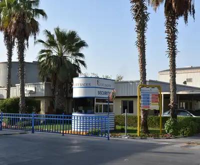 "Industrial facility entrance with security checkpoint and palm trees, Scott Bader manufacturing site for composites and resins" [G]