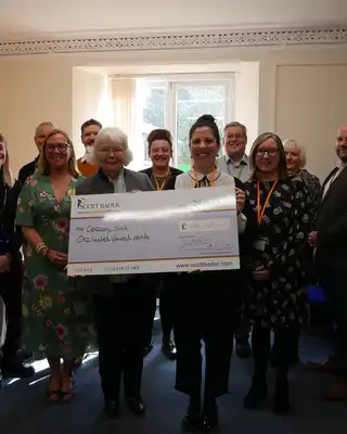 "Group presenting oversized Scott Bader check donation in bright meeting room with window view" [G]