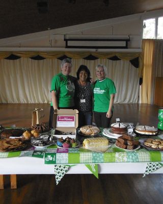 "Charity bake sale event with volunteers standing behind table of homemade desserts and refreshments in community hall" [G]
