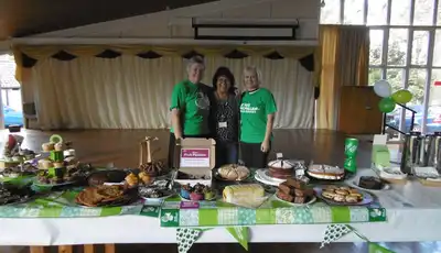 "Charity bake sale event with volunteers standing behind table of homemade desserts and refreshments in community hall" [G]