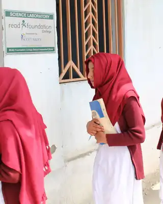 "Students in matching red headscarves and white uniforms walking through a school hallway carrying books and materials." [G]