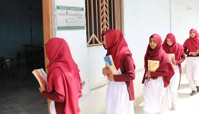 "Students in matching red headscarves and white uniforms walking through a school hallway carrying books and materials." [G]