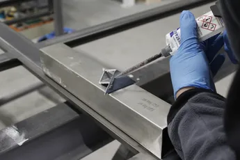 "Gloved hand applying Scott Bader structural adhesive to metal framework in manufacturing process" [G]