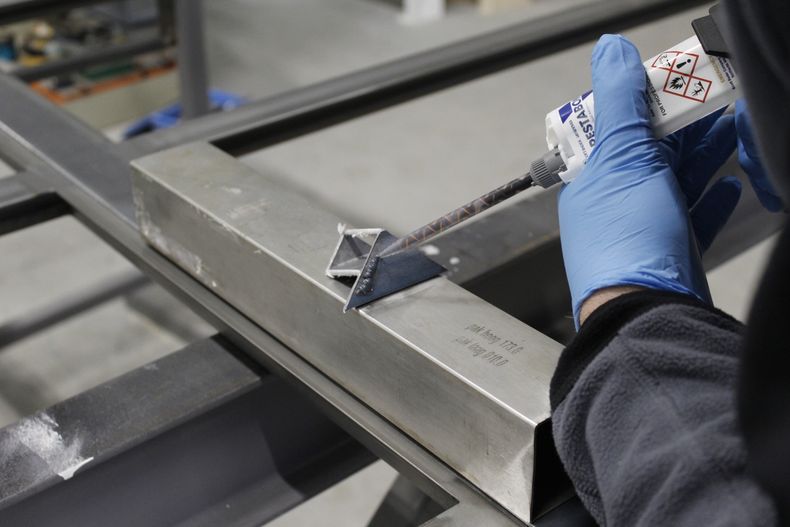 "Gloved hand applying Scott Bader structural adhesive to metal framework in manufacturing process" [G]