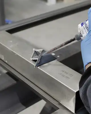 "Gloved hand applying Scott Bader structural adhesive to metal framework in manufacturing process" [G]
