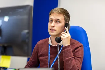 "Professional in a burgundy sweater speaking on phone at Scott Bader office with computer monitor visible" [G]