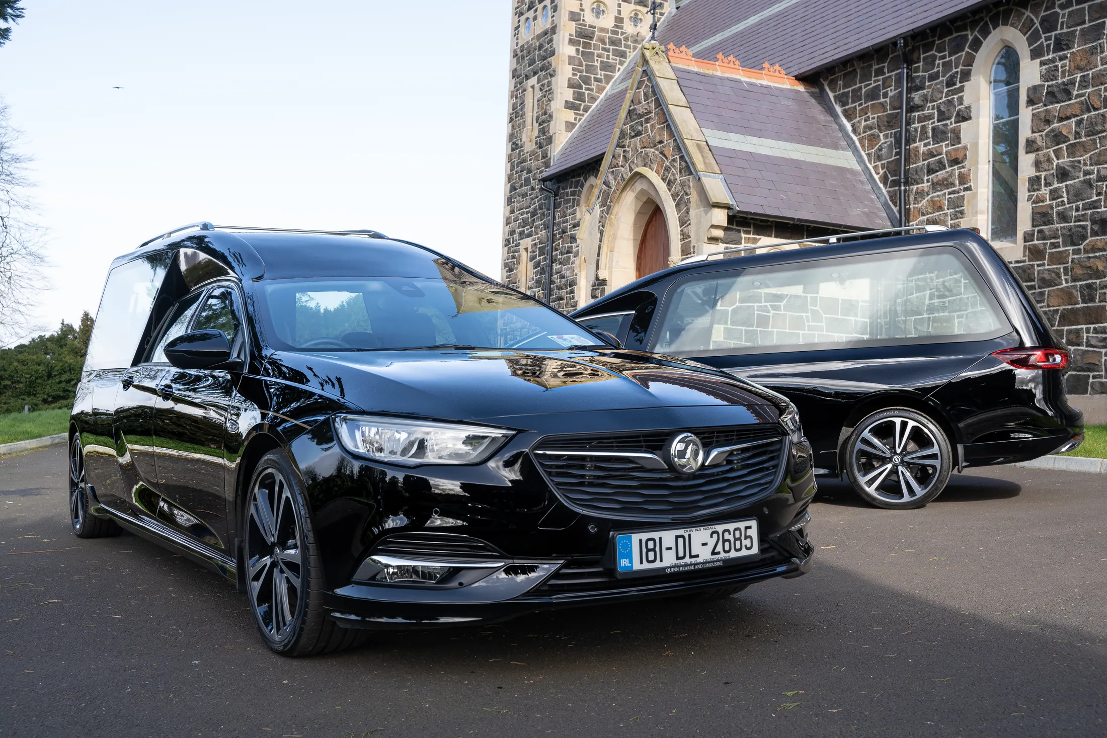 "Black hearse and funeral car parked outside stone church with arched entrance and stained glass windows" [G]