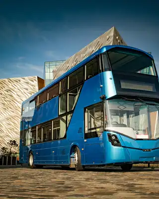 "Blue double-decker bus parked in front of the Titanic Belfast museum building with distinctive angular architecture" [G]