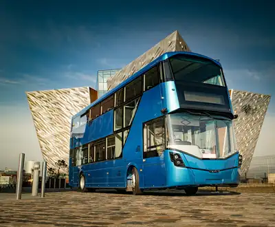 "Blue double-decker bus parked in front of the Titanic Belfast museum building with distinctive angular architecture" [G]