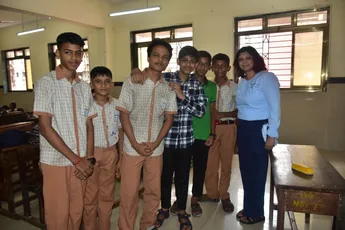 "Group of students and teacher posing together in a classroom with desks and windows visible in the background." [G]