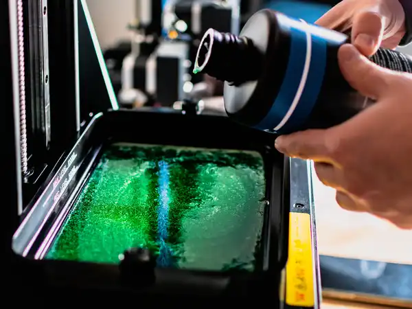 A person pouring liquid resin into a 3D printer vat filled with green liquid resin.