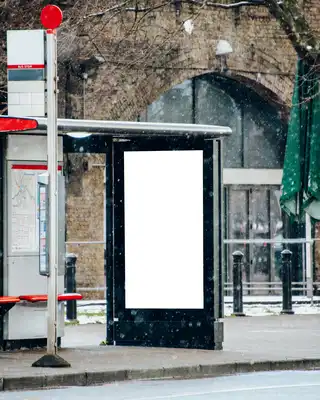 "Bus shelter with digital display panel in winter setting, snow-covered street with historic brick building in background" [G]