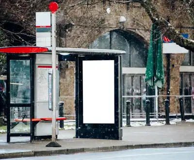 "Bus shelter with digital display panel in winter setting, snow-covered street with historic brick building in background" [G]