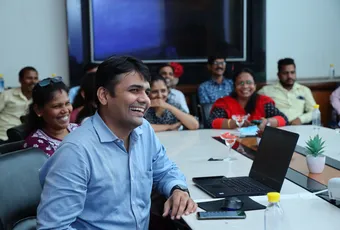 "Professional meeting in a conference room with attendees gathered around a table with laptops and presentation screen" [G]