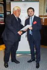 Two businessmen in suits exchanging documents at a formal event with a Commission banner in the background. [G]