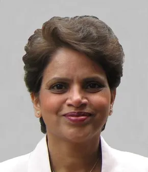 Professional headshot portrait of a woman in a white blazer against a grey background. [G]