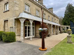 "Historic stone building with ornate porch and gravel pathway, featuring a modern sculpture on the lawn." [G]