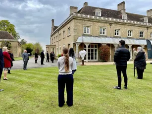 "Group gathering on lawn outside historic stone building at Scott Bader headquarters, showcasing the company's heritage." [G]