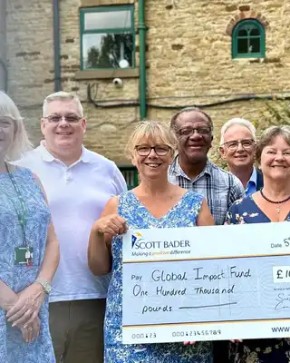 "Group of people standing outside a stone building, holding a large donation check with Scott Bader logo visible" [G]