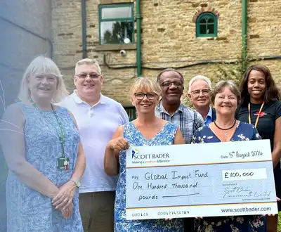 "Group of people standing outside a stone building, holding a large donation check with Scott Bader logo visible" [G]