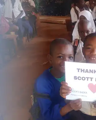 "Children in a classroom holding a 'Thank You Scott Bader' sign, showing community support for the company's initiatives." [G]