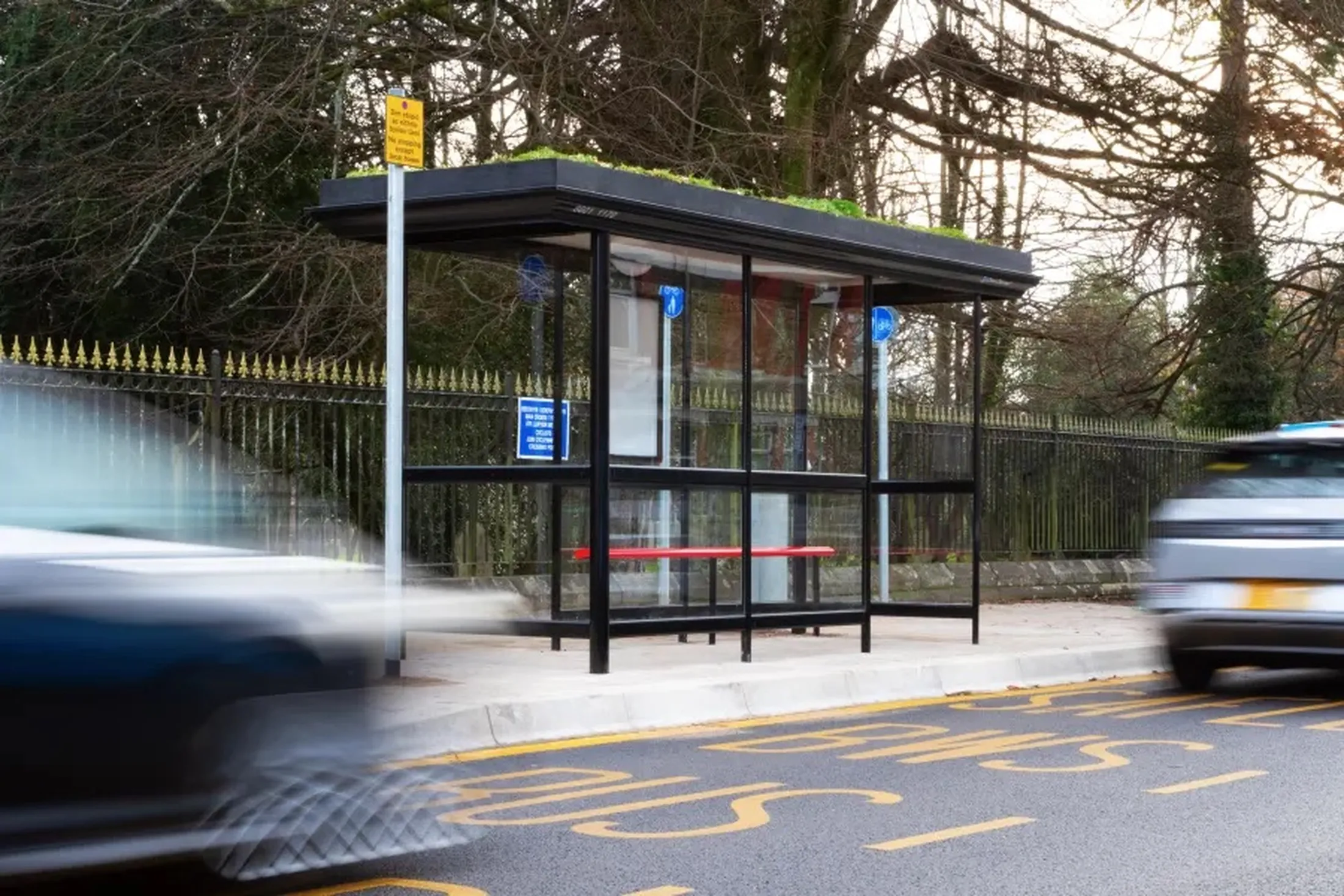 "Bus shelter with motion-blurred vehicles passing by on a roadside, featuring a bench and information panel." [G]