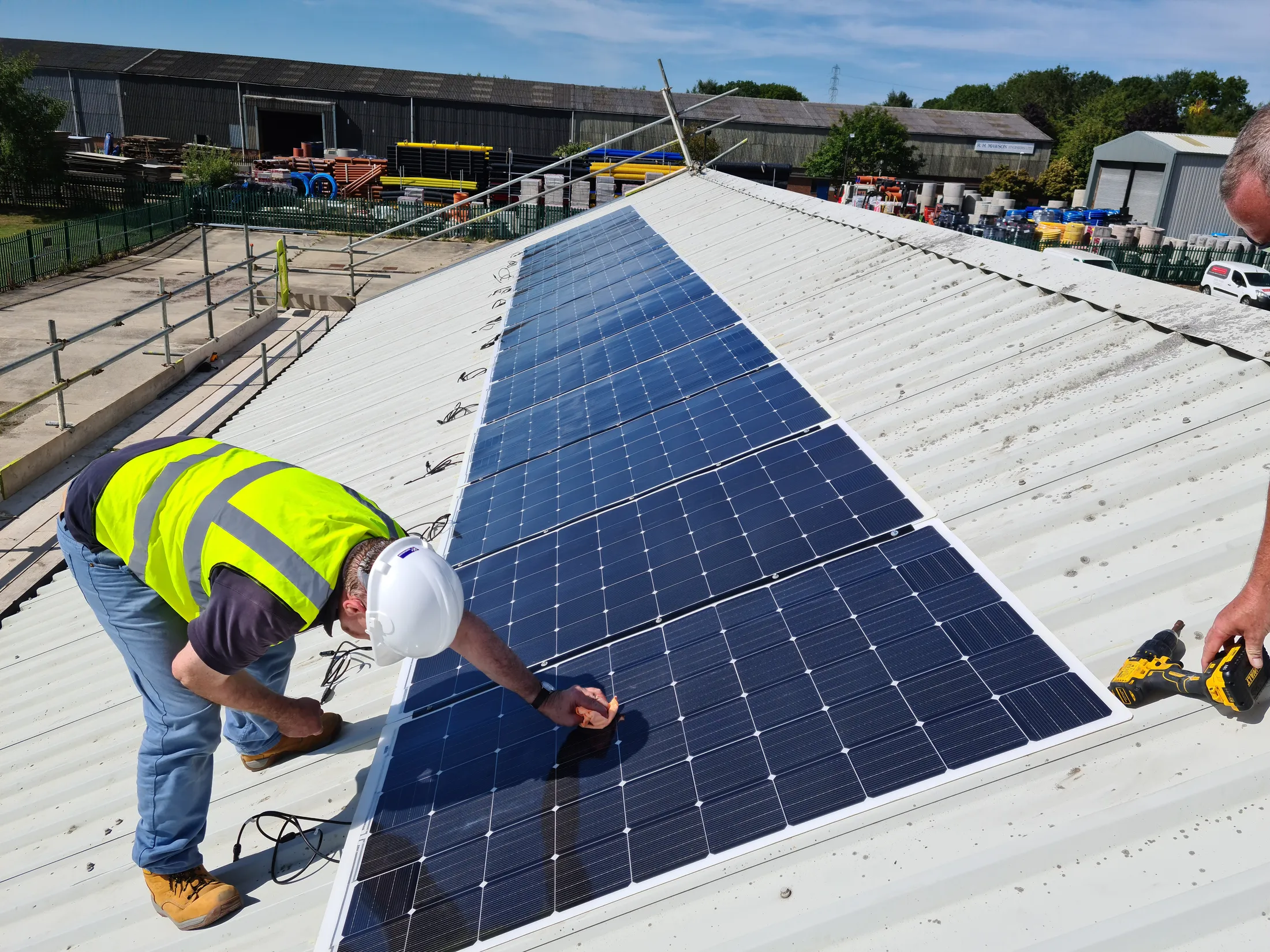 "Technician installing solar panels on industrial roof at Scott Bader facility with composites manufacturing buildings visible in background" [G]