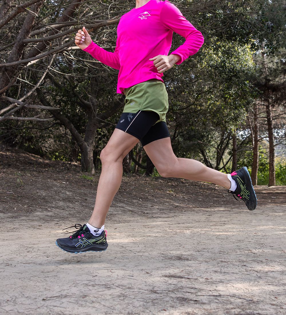 A person in a pink long-sleeved shirt and green shorts jogging on a dirt path surrounded by trees.