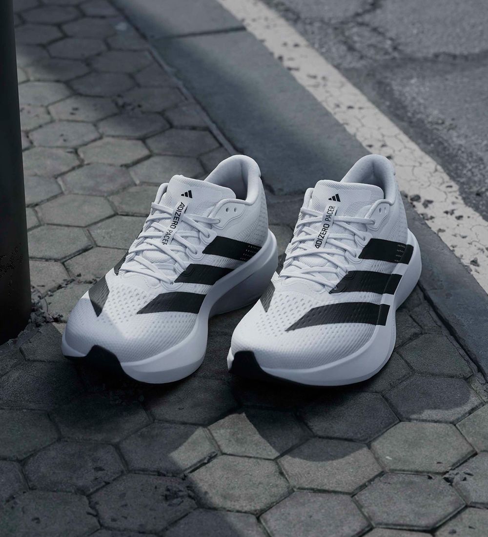 White sports shoes with black stripes placed on a hexagonal tile sidewalk near a street curb.