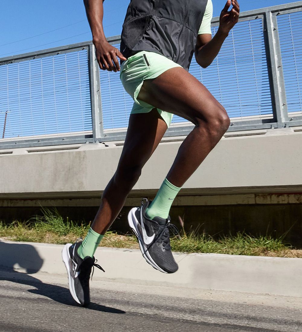 A person in green shorts and socks runs along a road, wearing black and white athletic shoes near a fence under clear blue skies.