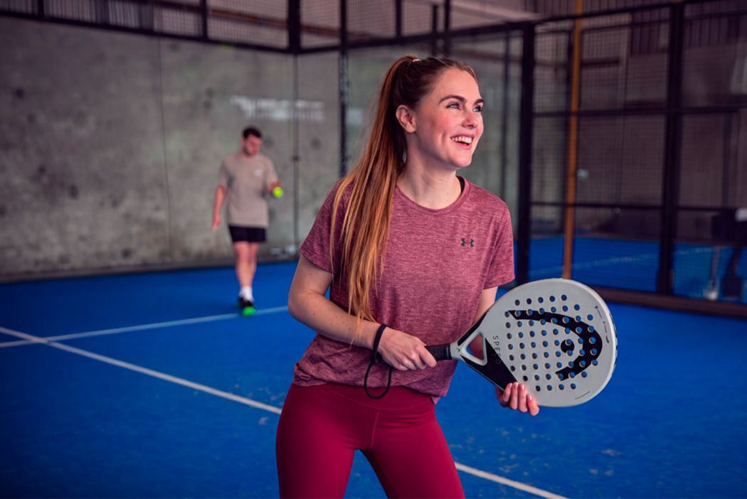 A woman in athletic gear holds a padel racket and smiles on a blue court, while a person in the background prepares to play.