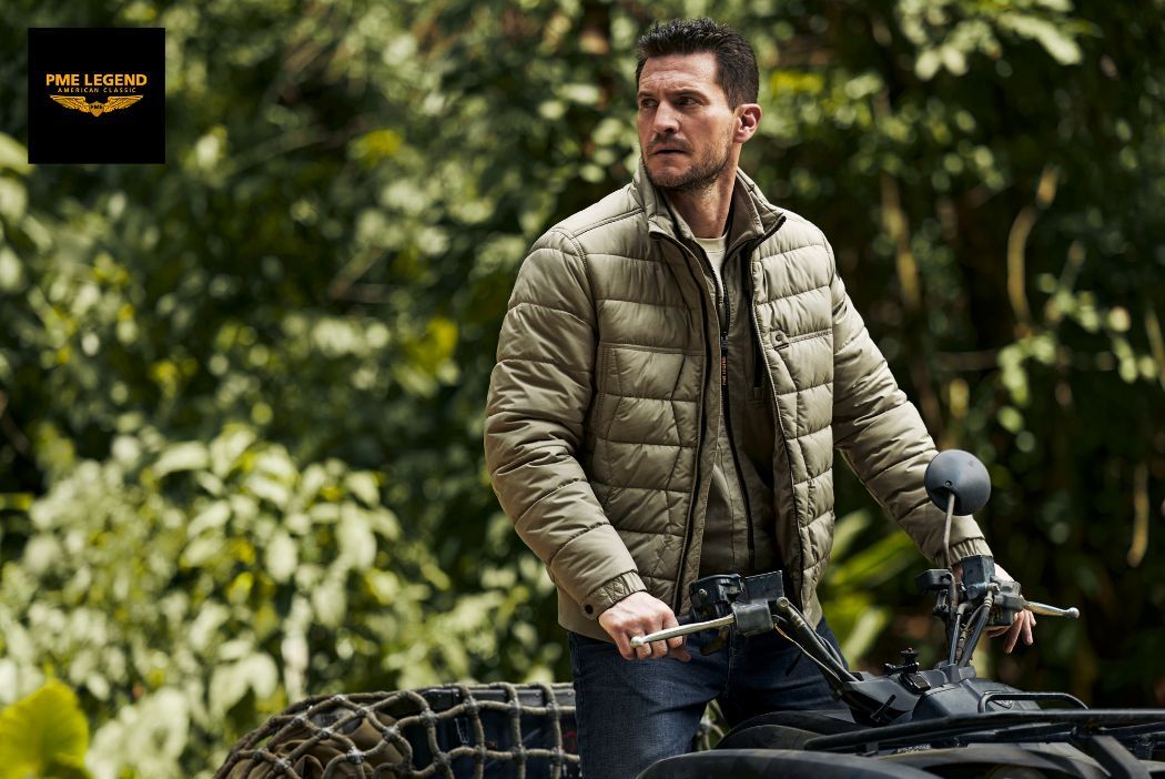 A man in a green quilted jacket stands with a motorcycle in a lush green forest, with the PME Legend logo in the top left corner.