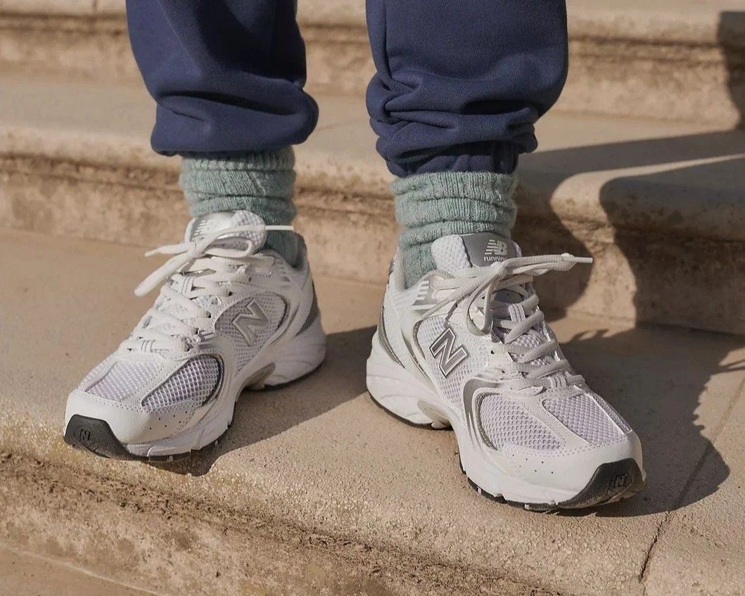White New Balance sneakers with gray accents on a person wearing blue pants and green socks, standing on beige stone steps.
