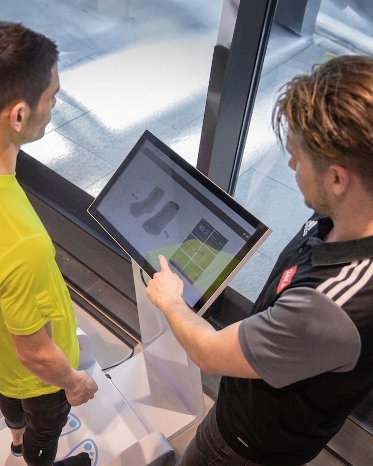 A person in a yellow shirt stands on a foot scanner while another person points at a touchscreen displaying foot measurements.