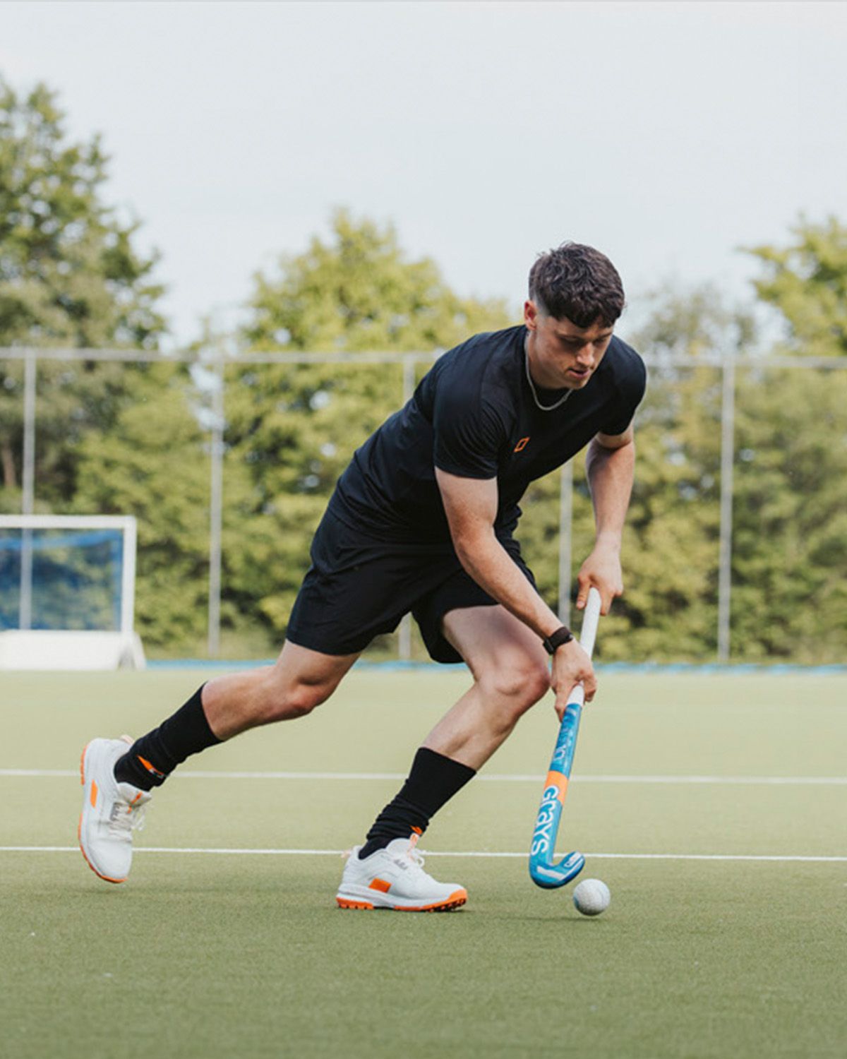 A man in black sports attire plays field hockey, focusing intently as he controls the ball with his stick on an outdoor field.
