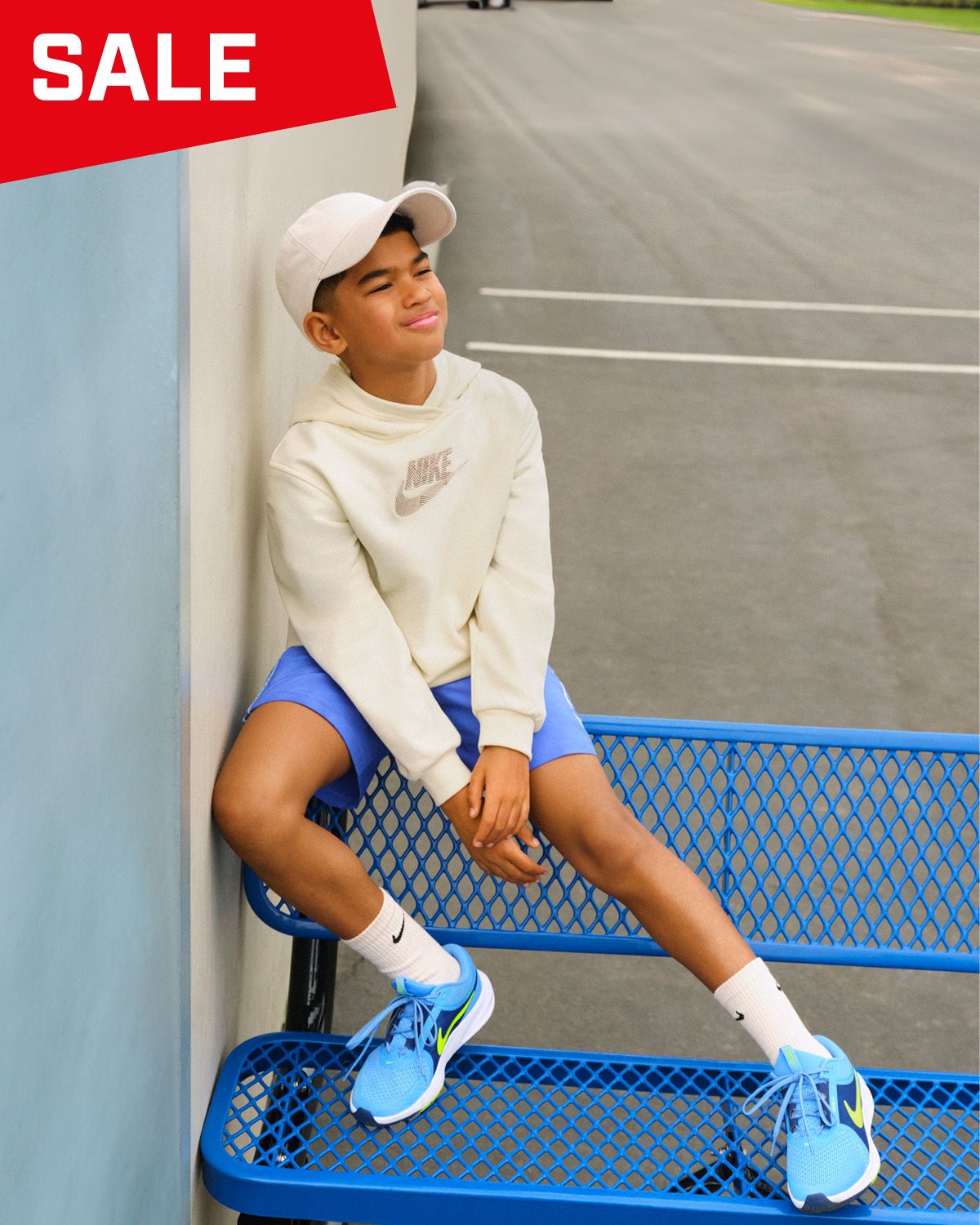 Young person in a white hoodie and blue shorts sitting on a blue bench, wearing blue sneakers and a white cap. "Sale" text in red at the top.