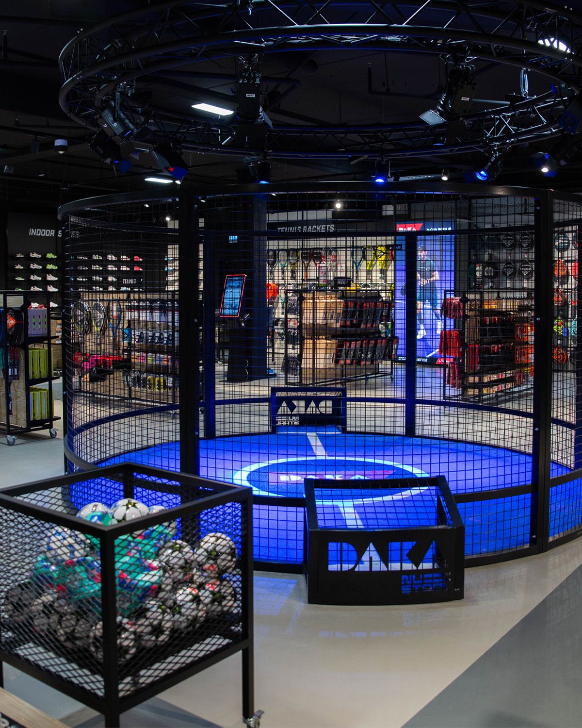 Indoor sports store with blue caged area, overhead lighting, various equipment displays, and a cart filled with soccer balls in the foreground.
