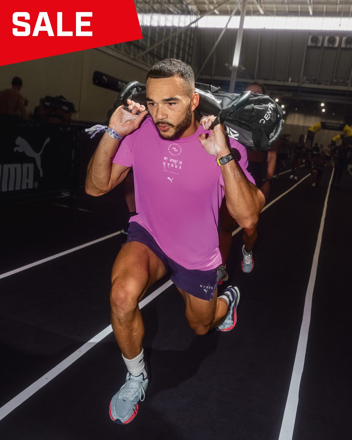 Athlete in pink shirt and shorts performing a sandbag lunge indoors, with a prominent "SALE" sign in the corner.