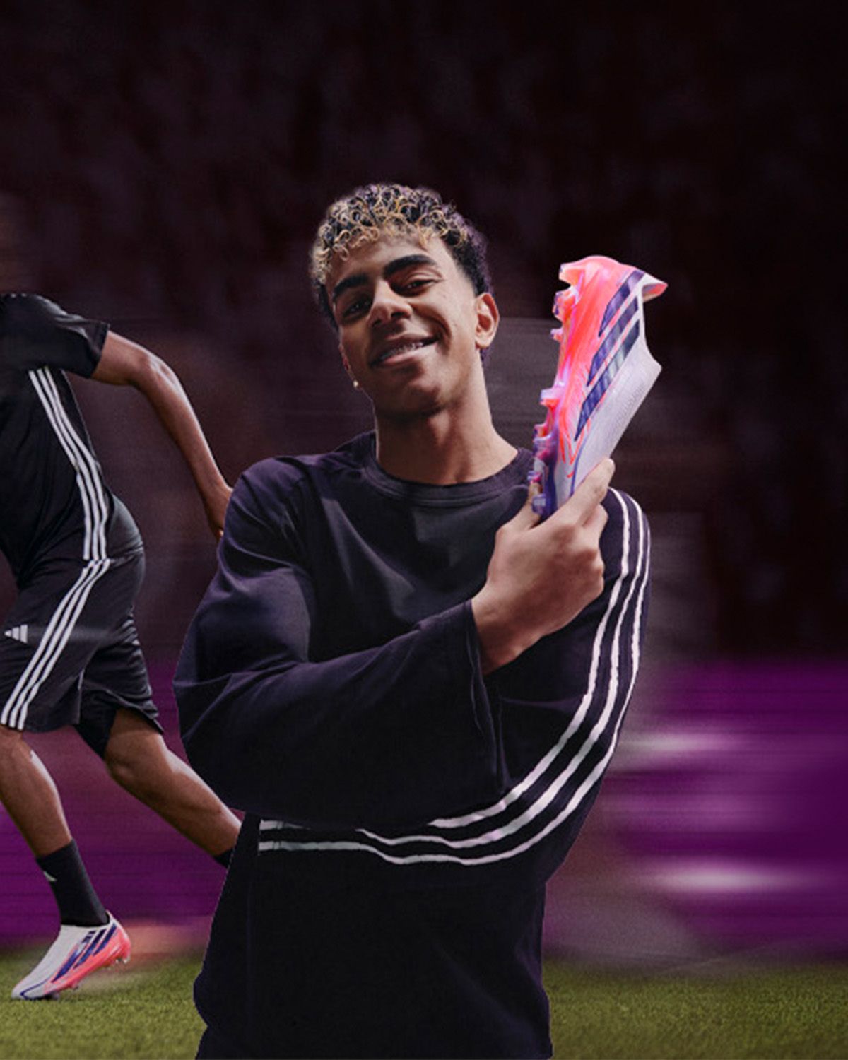A smiling person in a black outfit holds a vibrant soccer shoe with pink and blue accents, with another blurred figure in the background.