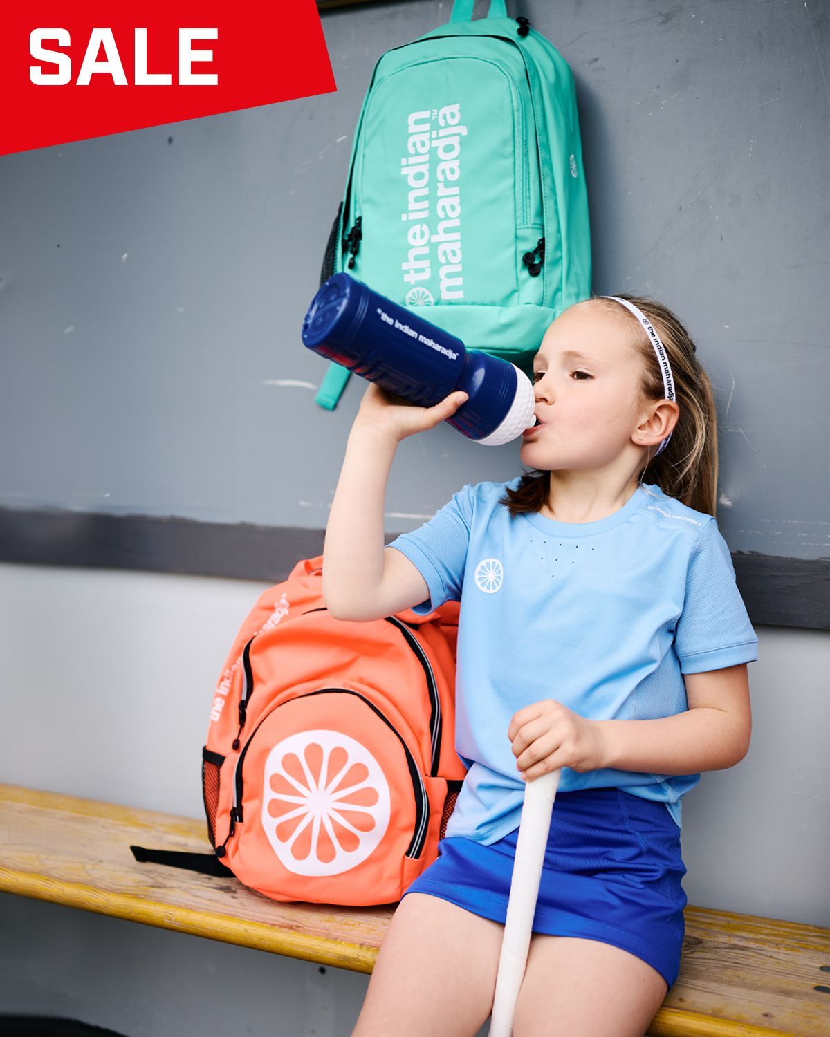 Young girl in sports attire drinking from a bottle, sitting on a bench, with two backpacks. A "SALE" sign is displayed at the top corner.