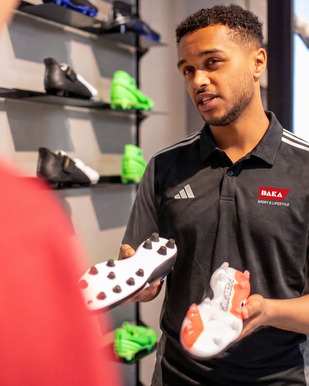 Store employee in a black polo showing soccer cleats to a customer, with a display of various colorful cleats in the background.