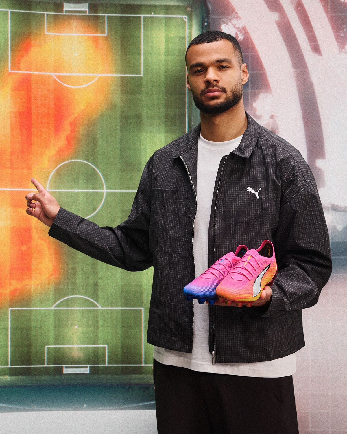 Man holding pink soccer cleats, standing in front of a colorful soccer field heat map, wearing a black Puma jacket.